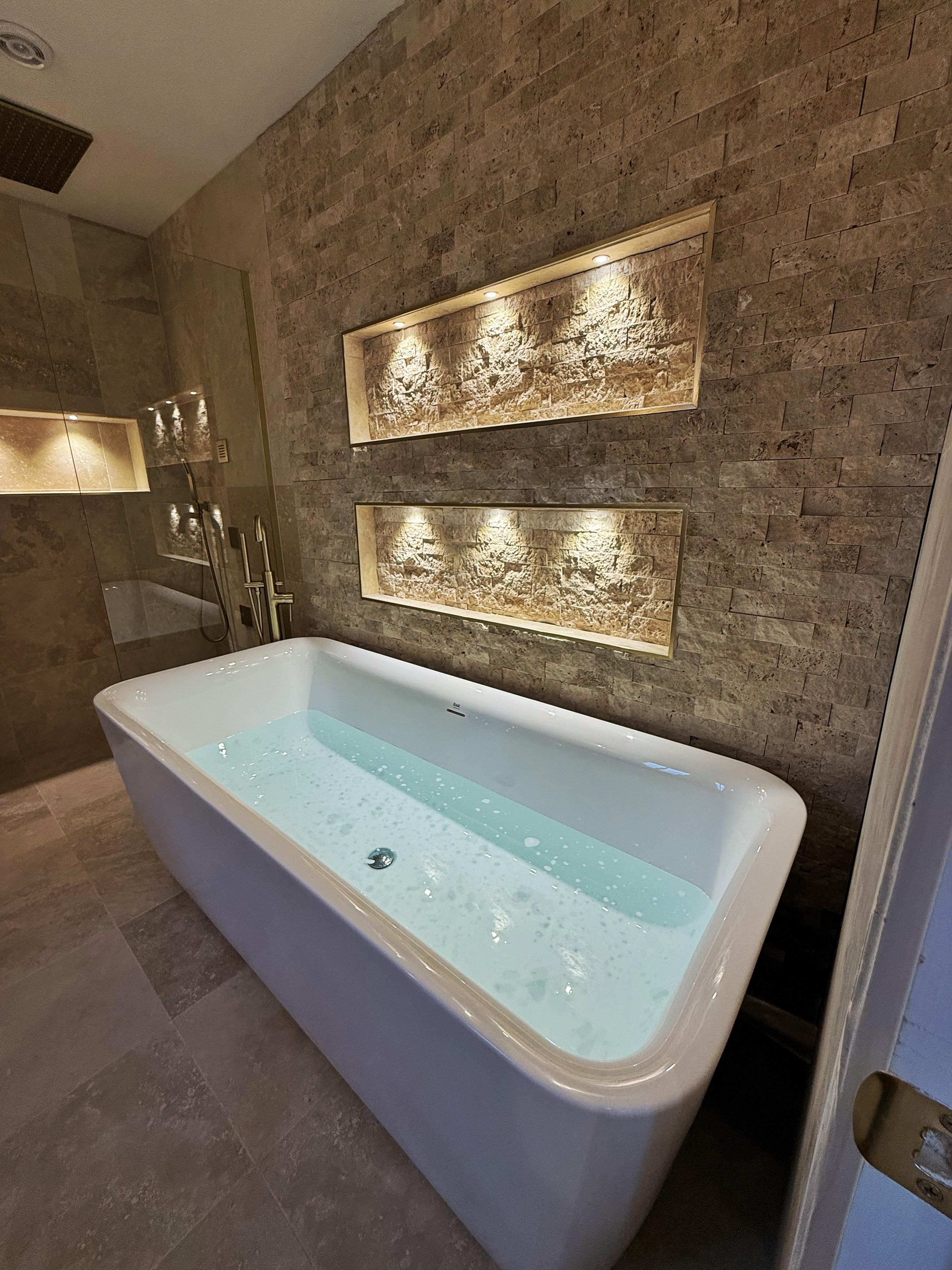 Freestanding bath in a stone-tiled bathroom with lit recessed alcoves