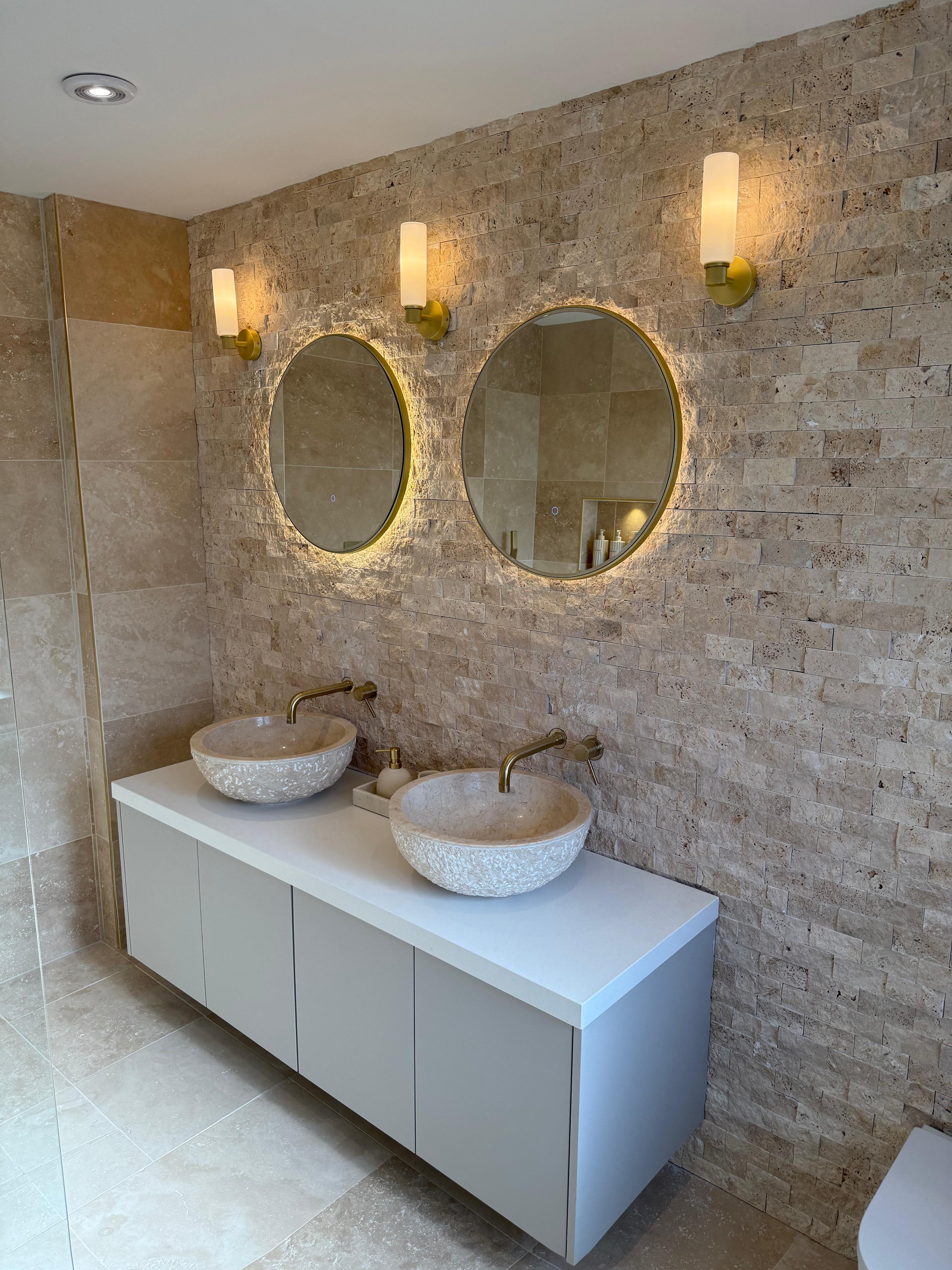 Stone-tiled bathroom with twin vessel basins, round backlit mirrors and brass wall lights