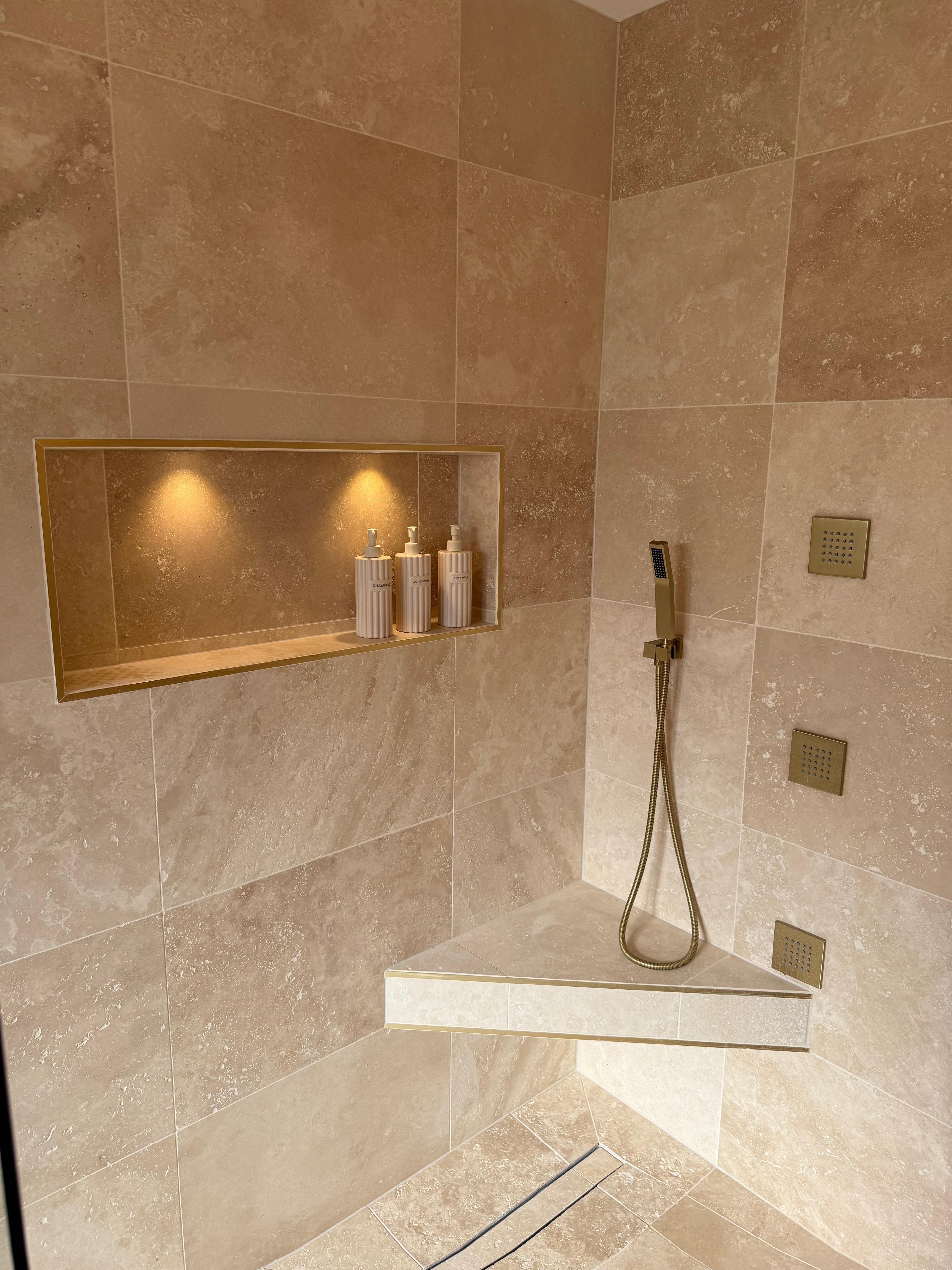 Walk-in stone shower with a corner seat, brass controls and a lit niche